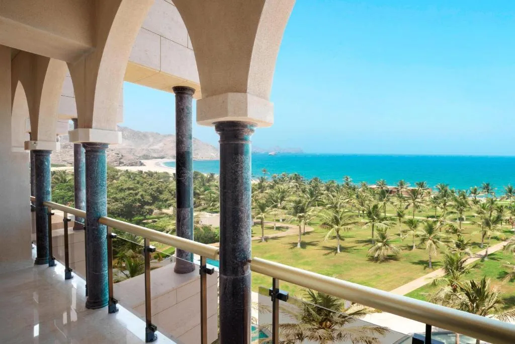 A balcony view features palm trees on a grassy area, leading to a sandy beach and blue ocean. The scene is framed by archways and columns. It's bright and sunny.