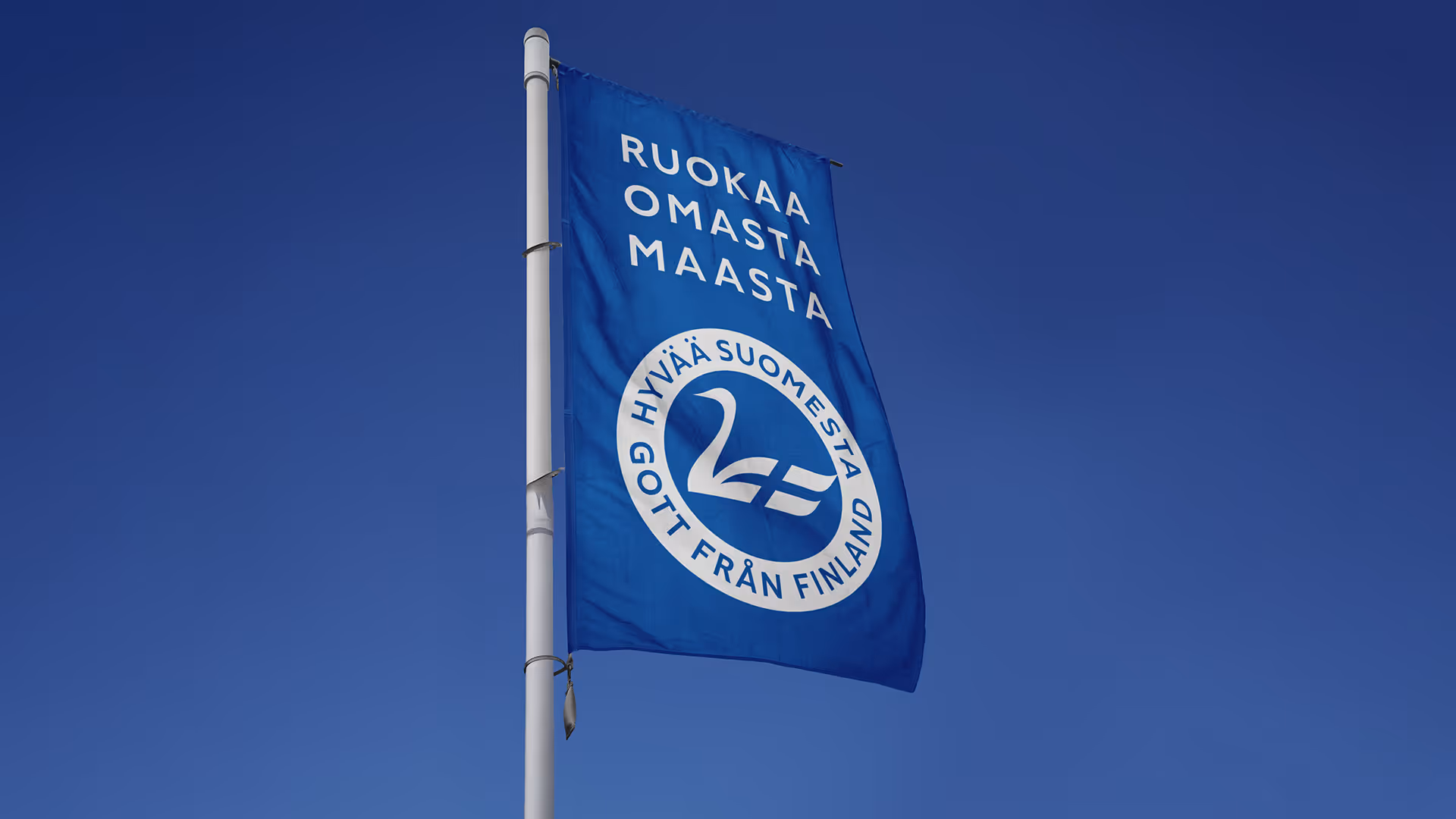 Liehuva Hyvää Suomesta -lippu lipputangossa sinistä taivasta vasten.