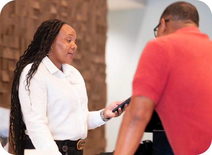 Hotel front desk employee speaking with a guest at the reception desk while holding a mobile device.
