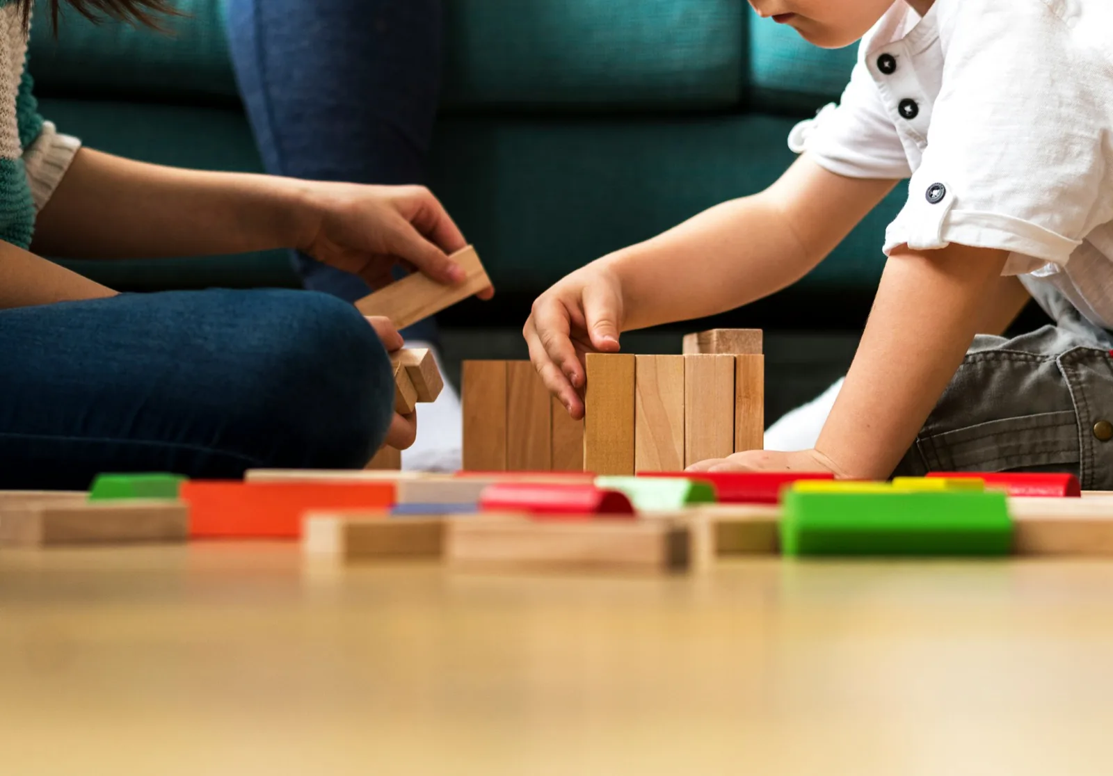Mains d'une adulte et d'un enfant construisant une tour de blocs en bois au sol.