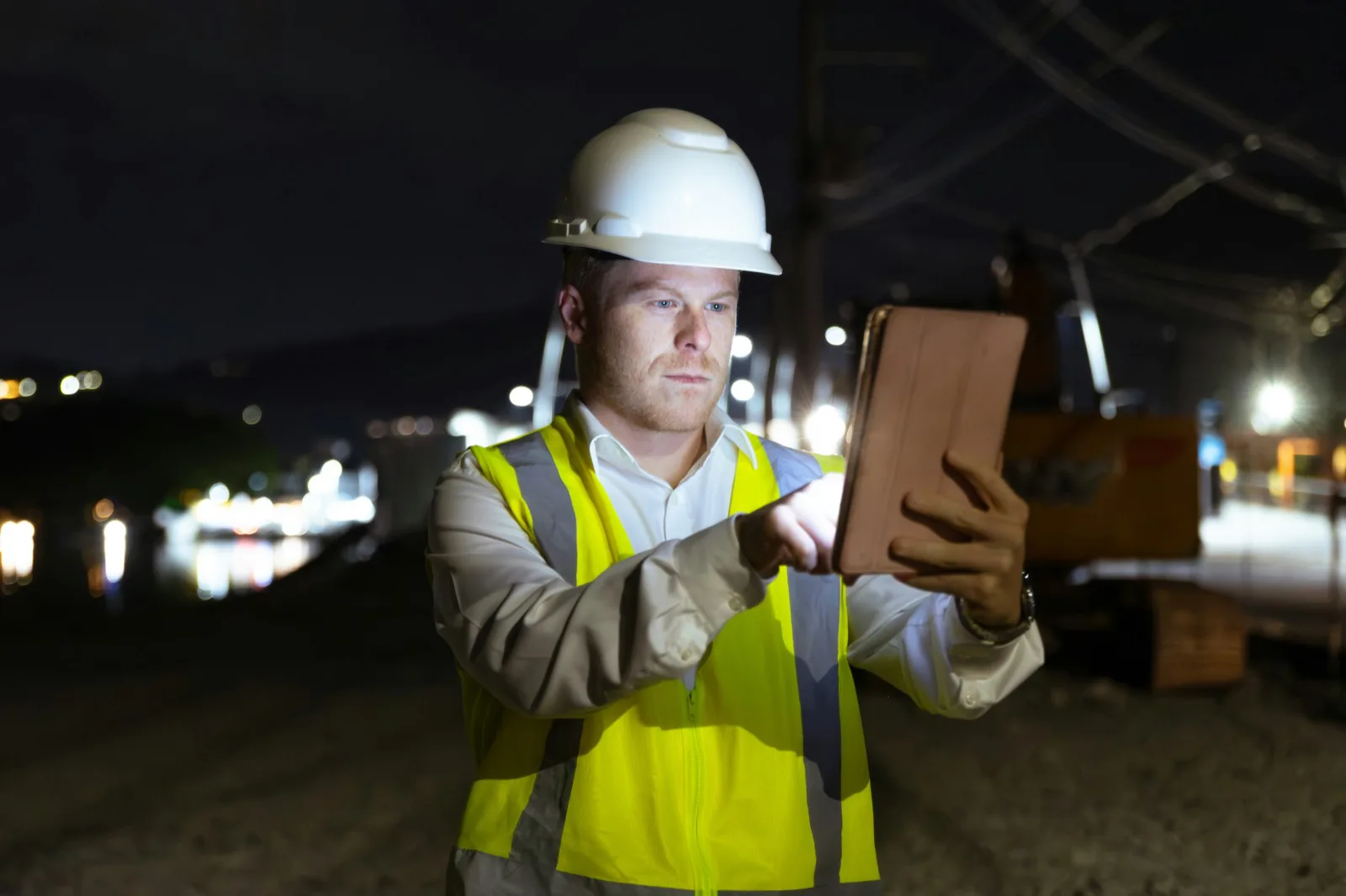 Un technicien en casque et gilet haute visibilité consulte une tablette de nuit sur un chantier ferroviaire.