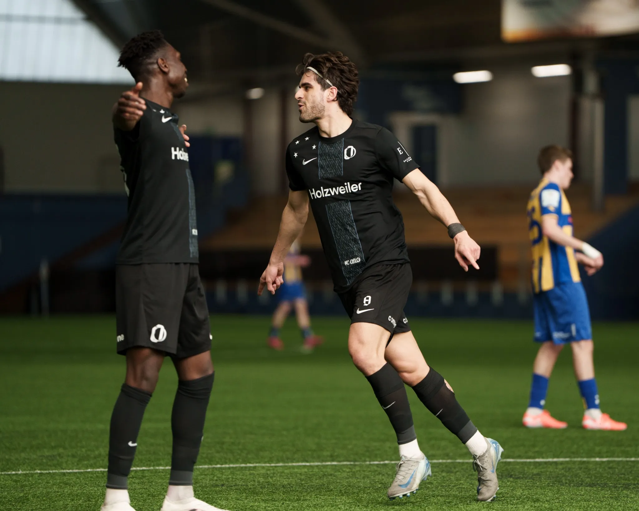 To fotballspillere i svarte drakter jubler på kunstgressbane innendørs, mens en spiller i blått og gult går i bakgrunnen.