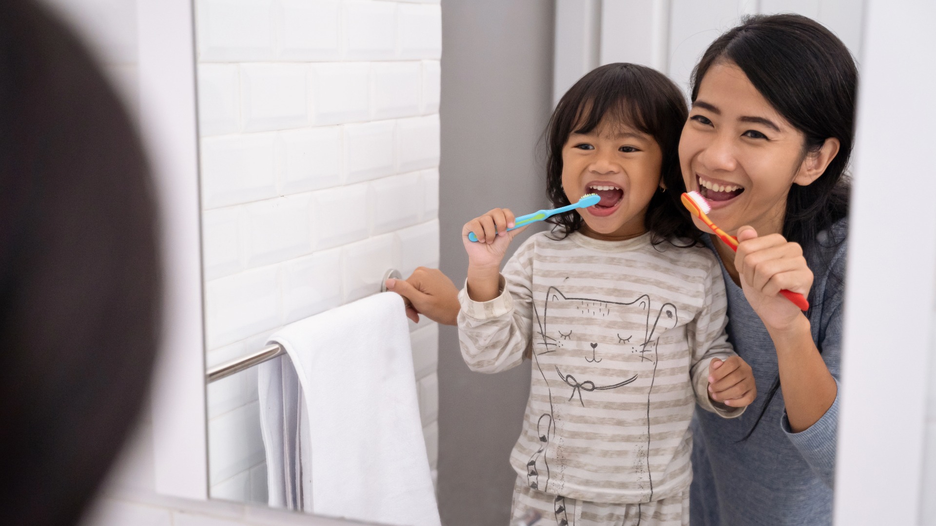 A mother and child brushing their teeth together in the bathroom mirror