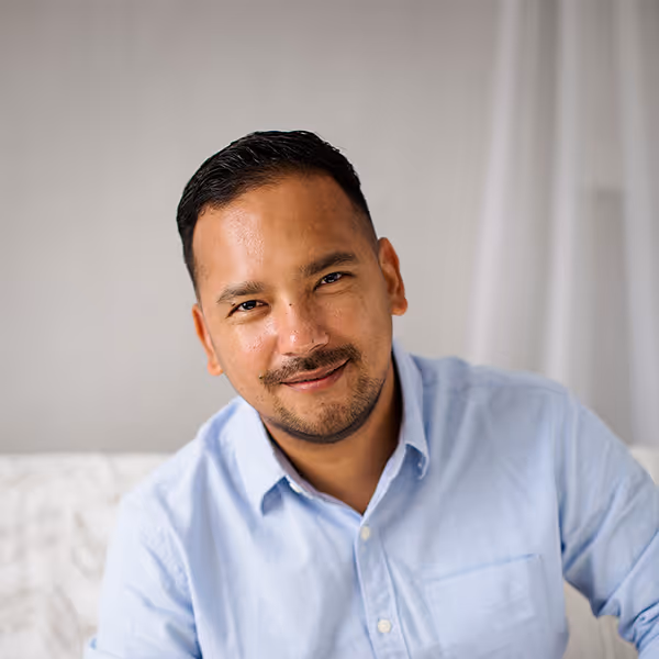 Smiling man with short dark hair and a mustache wearing a light blue button-up shirt sitting indoors. - Muserna