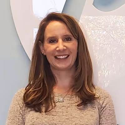 Smiling woman with long brown hair wearing a beige sweater and necklace, standing indoors.