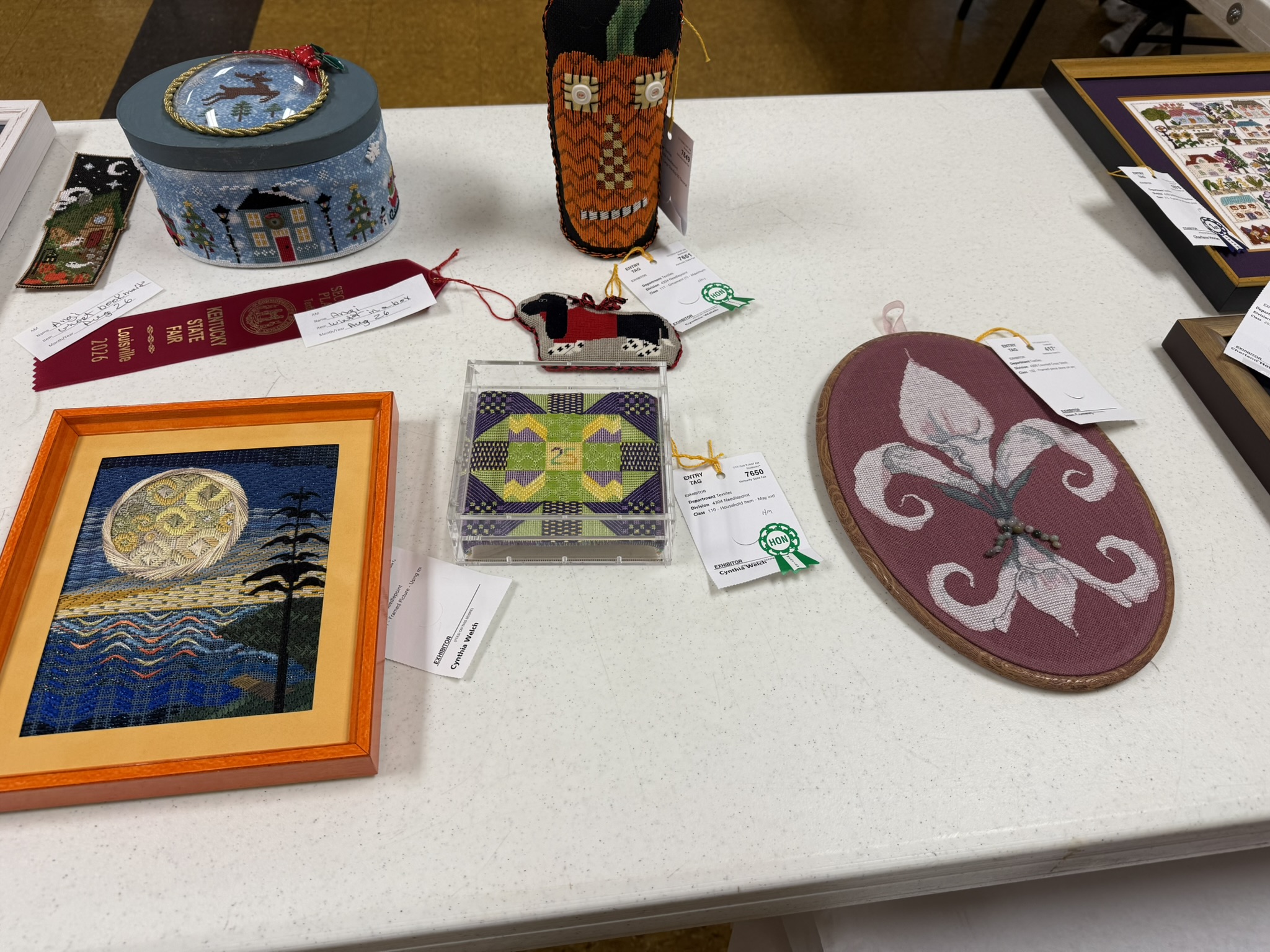 Table of small stitched pieces including a decorated box, a framed moon scene and a fleur-de-lis hoop