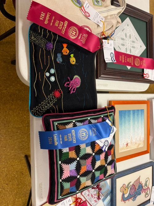 Entries on a display table including a black underwater embroidery and a log cabin quilt square with ribbons