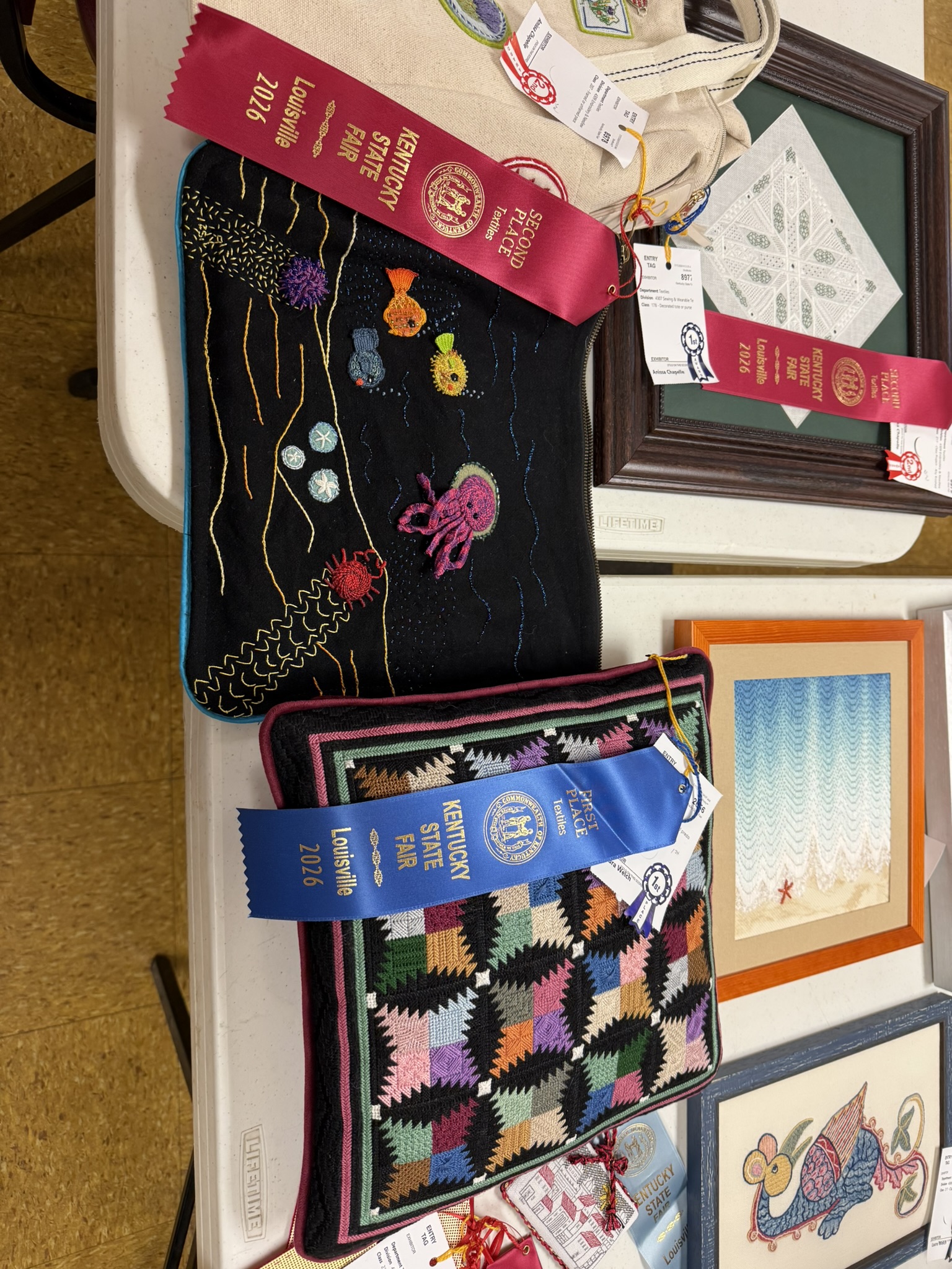 Entries on a display table including a black underwater embroidery and a log cabin quilt square with ribbons