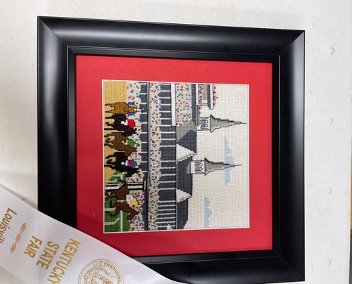 Framed cross stitch of the Churchill Downs twin spires and a race crowd, with a Kentucky State Fair ribbon