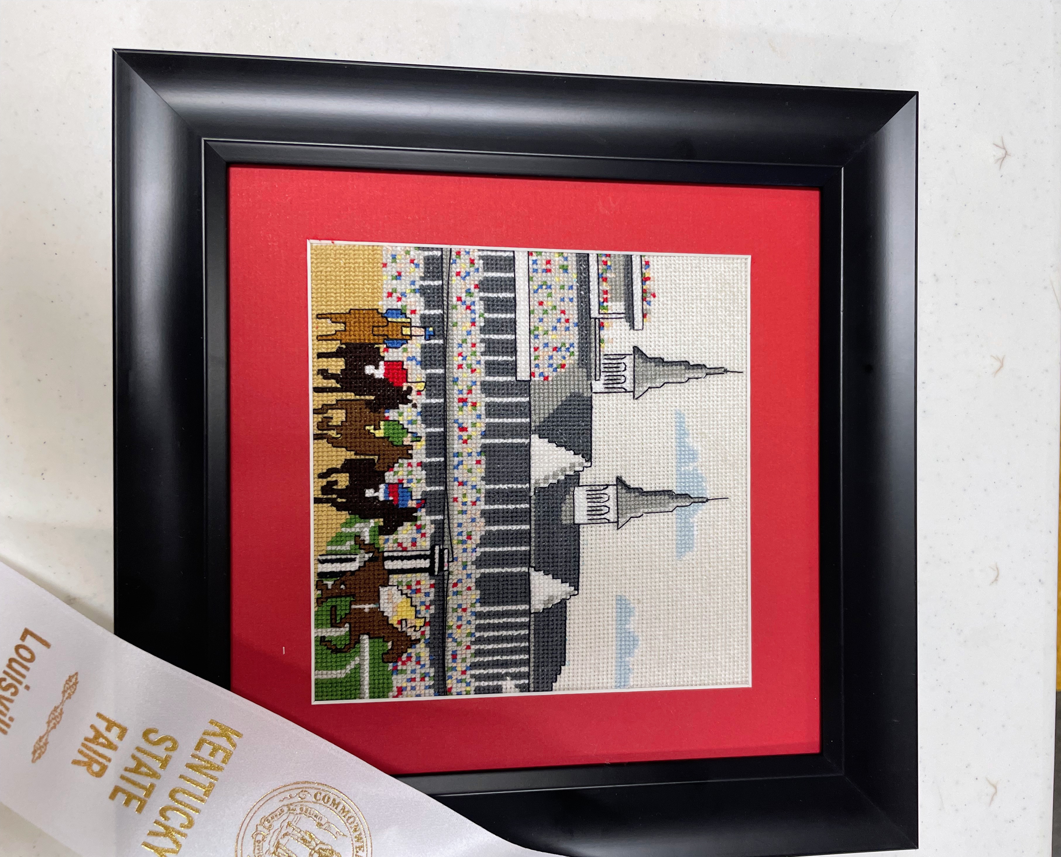 Framed cross stitch of the Churchill Downs twin spires and a race crowd, with a Kentucky State Fair ribbon