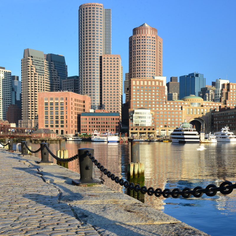 Boston waterfront with cobblestone walkway, black chain fence, and city skyline in the background.