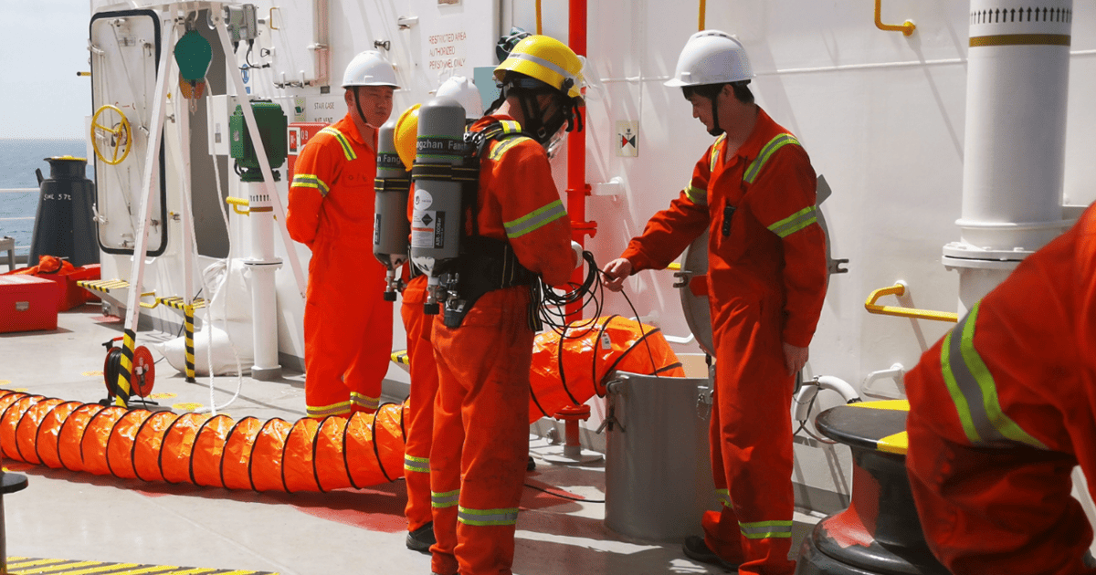 Offshore workers in orange safety suits and helmets coordinate on a ship’s deck beside hoses and equipment, conveying teamwork and readiness.