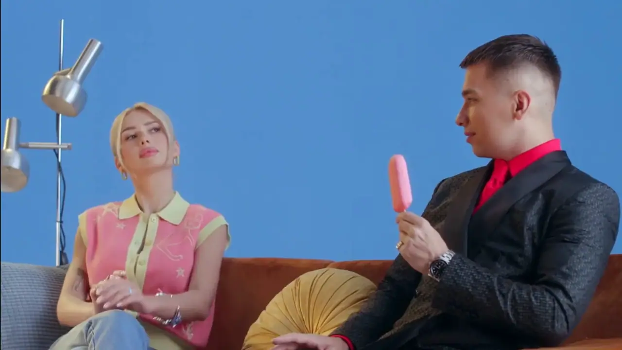 Two actors on a sofa holding pink ice lollies, a blue set behind them on the LED wall