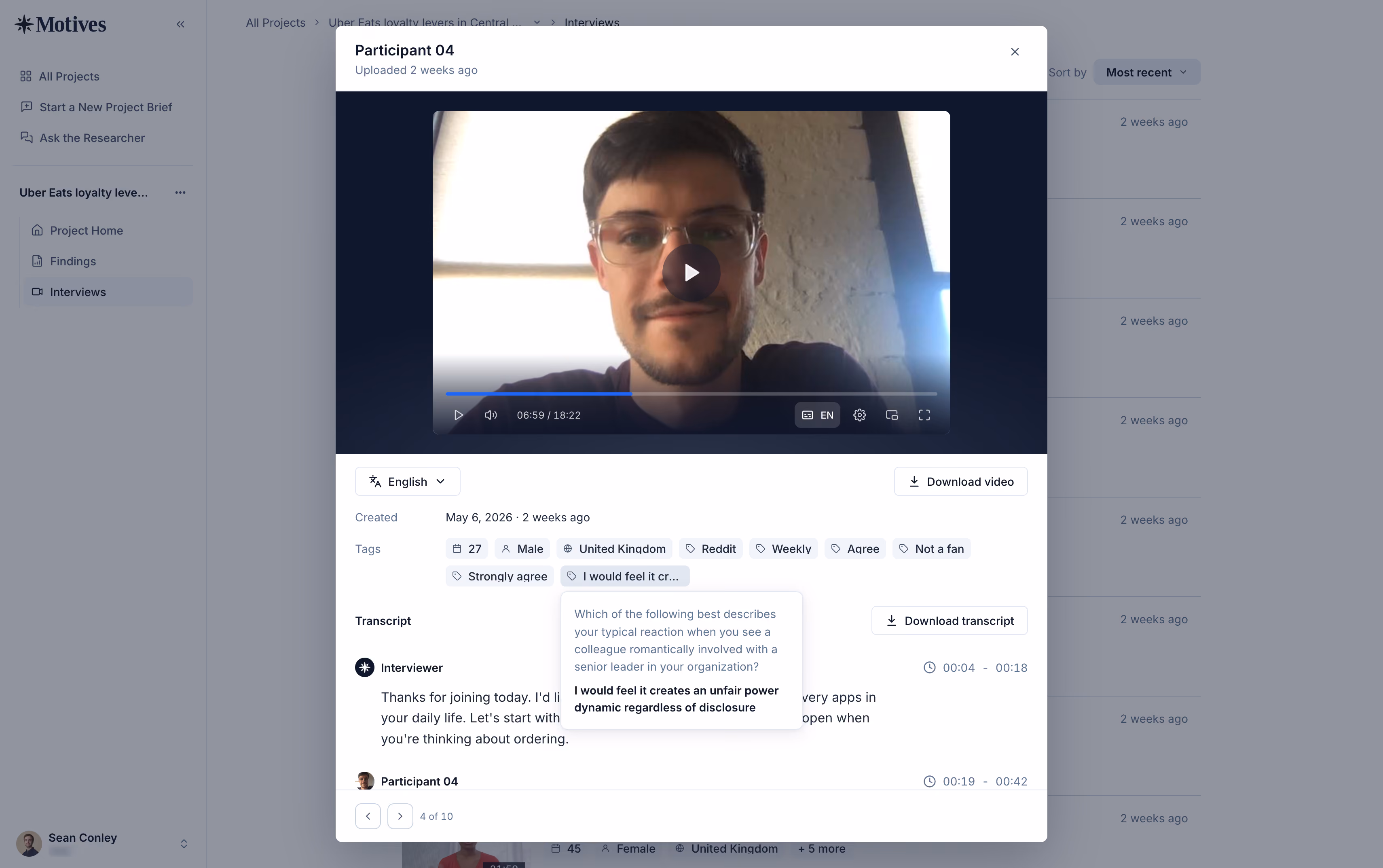 Screenshot of an online interview platform showing a video interview with Participant 04, a young man with short brown hair and glasses, sitting in front of a window. The video is paused at 6:59 of 18:22. Below the video, metadata shows participant details: age 27, male, United Kingdom, Reddit user, weekly app usage, opinions tagged as Agree, Not a fan, Strongly agree. A transcript section shows the interviewer asking about the participant's reaction to a colleague romantically involved with a senior leader, and a response highlighting unfair power dynamics. Controls for language, downloading video and transcript are visible.
