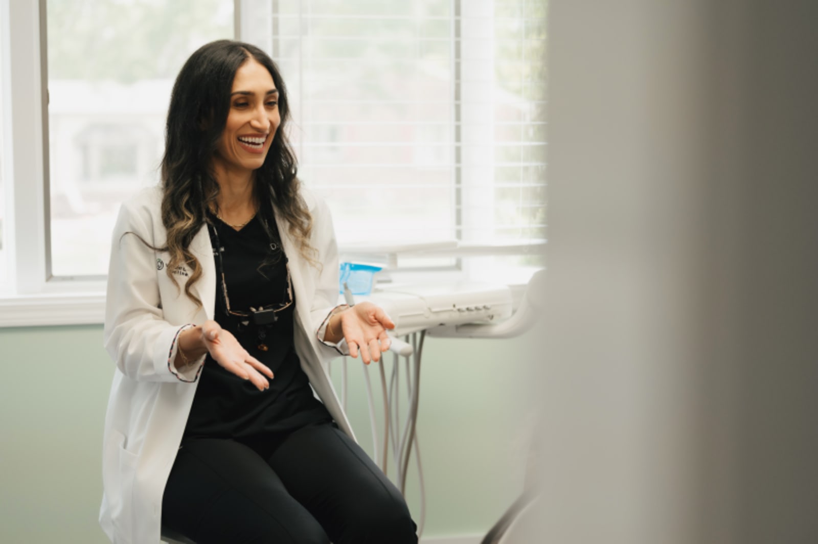 Female dentist in white coat gesturing while speaking in a modern dental office treatment room.
