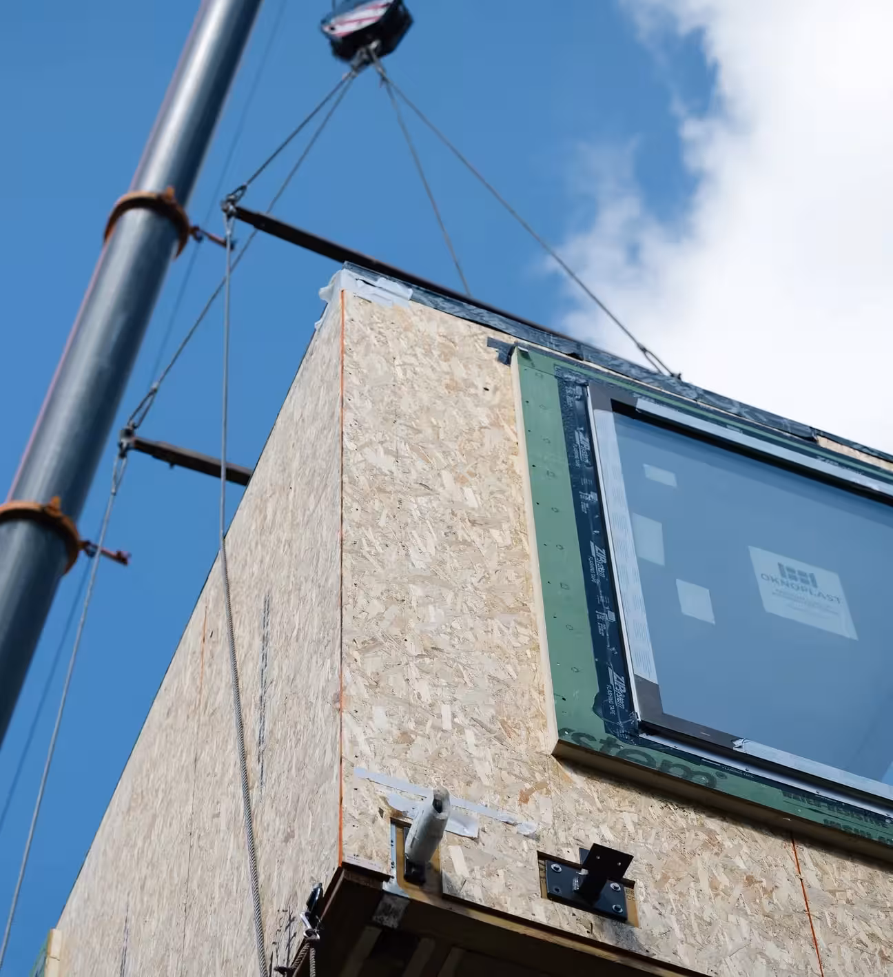 Wooden building section with a window being lifted by crane cables against a blue sky.