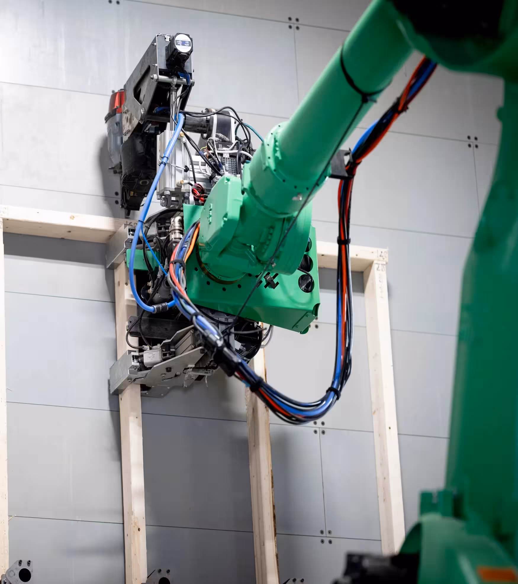 Green robotic arm with wires attached to a metal and wooden wall frame in an industrial setting.