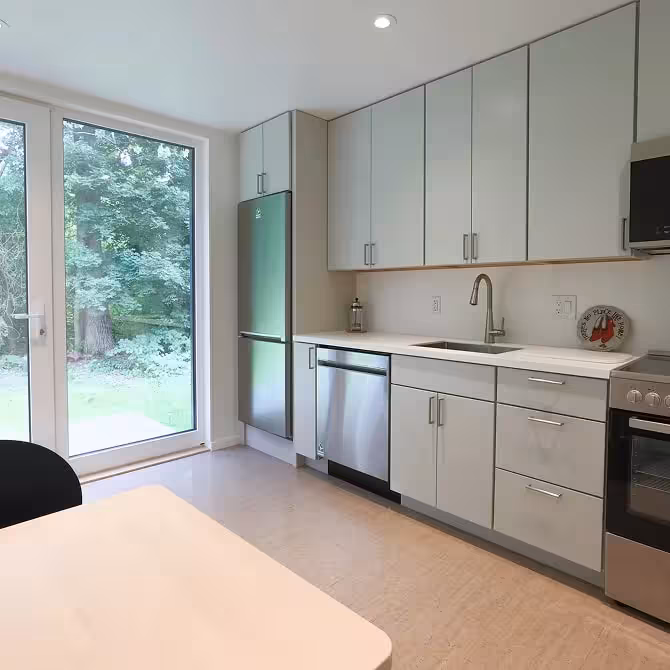 Modern kitchen with white cabinets, stainless steel appliances, and a large glass door showing greenery outside.