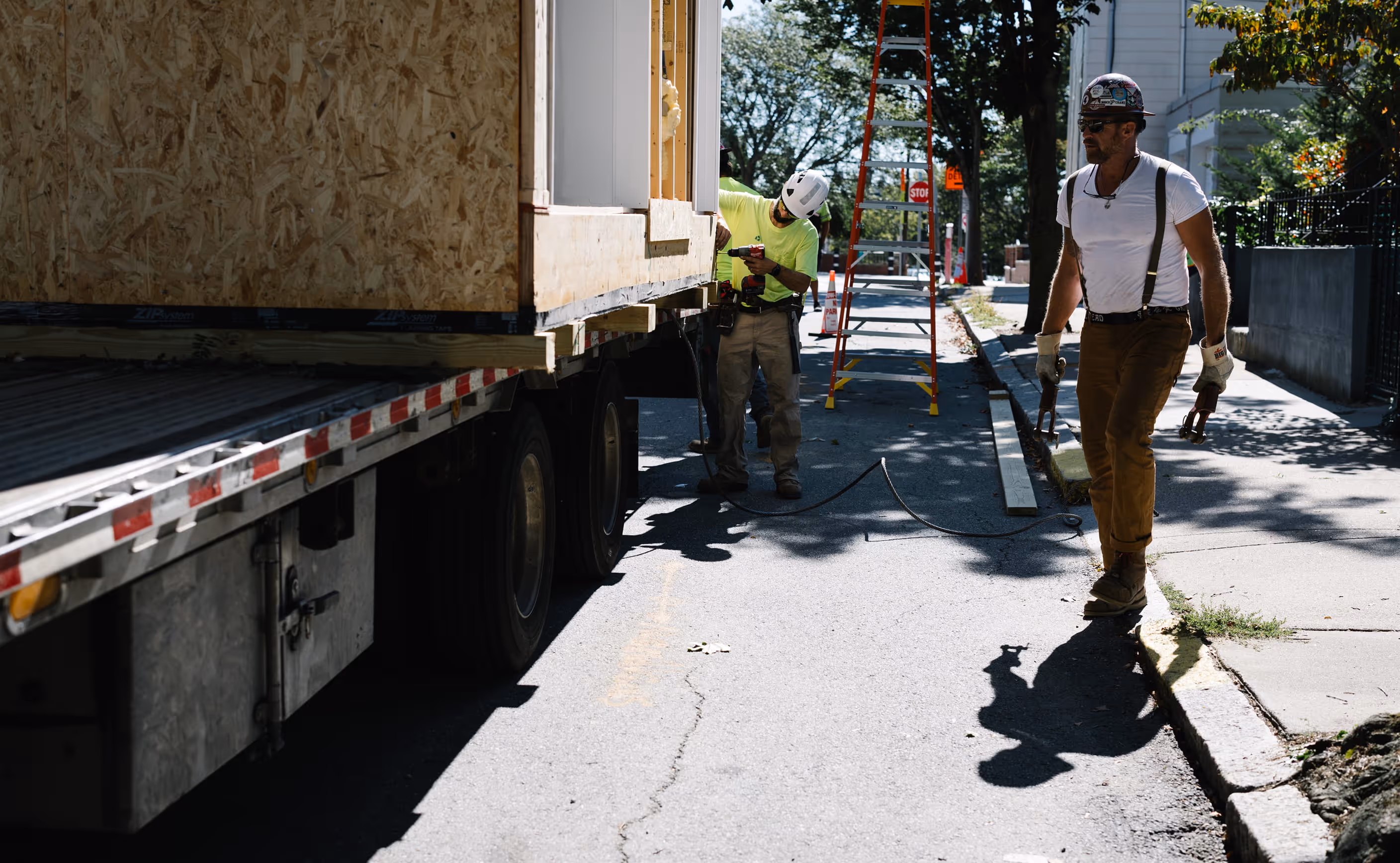 Two construction workers near a flatbed truck unloading a wooden structure on a sunny street.
