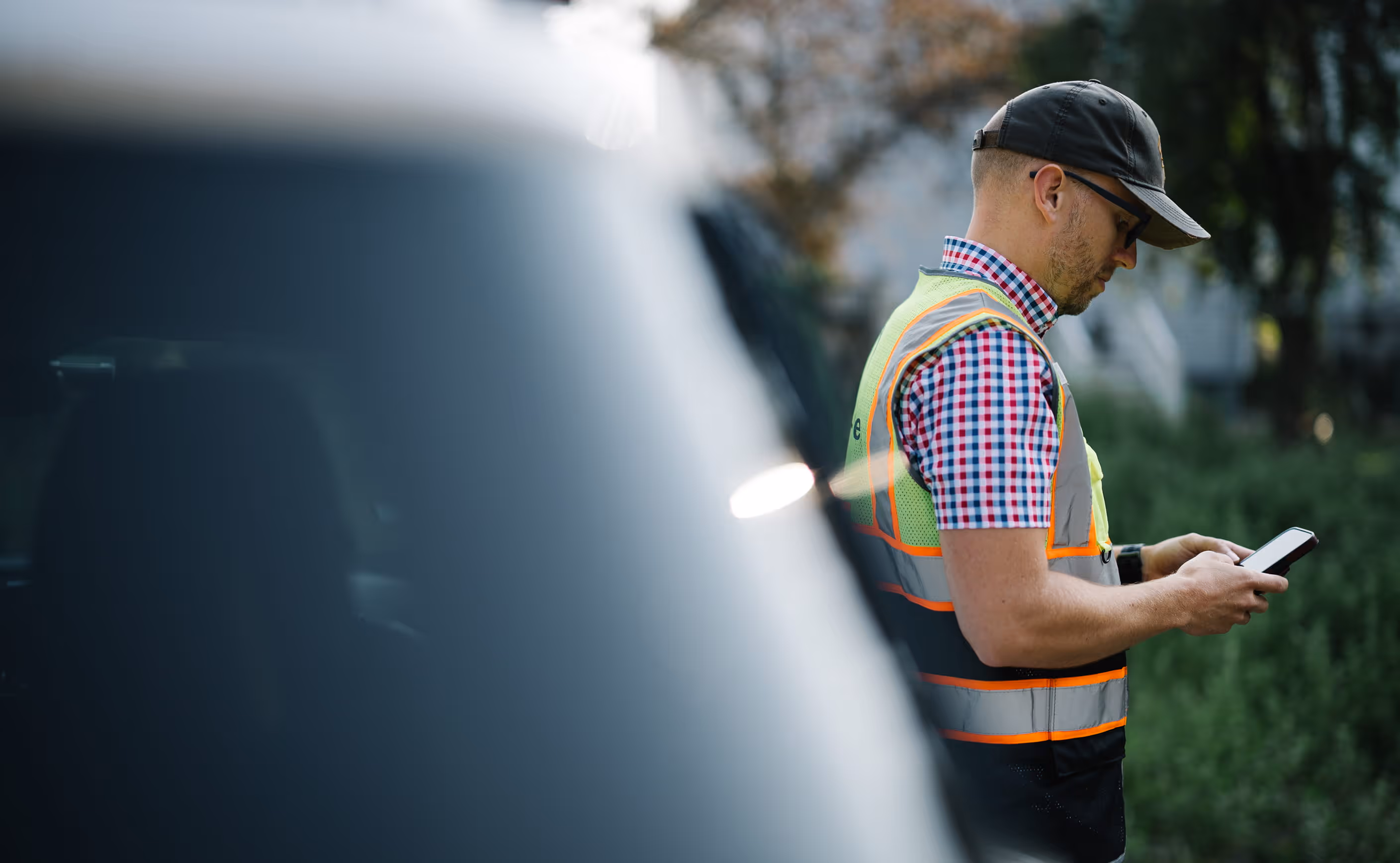 Man wearing safety vest and cap looking at smartphone outdoors beside a blurred vehicle.
