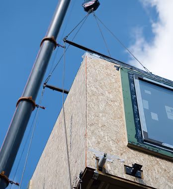 Wooden building section with a window being lifted by crane cables against a blue sky.