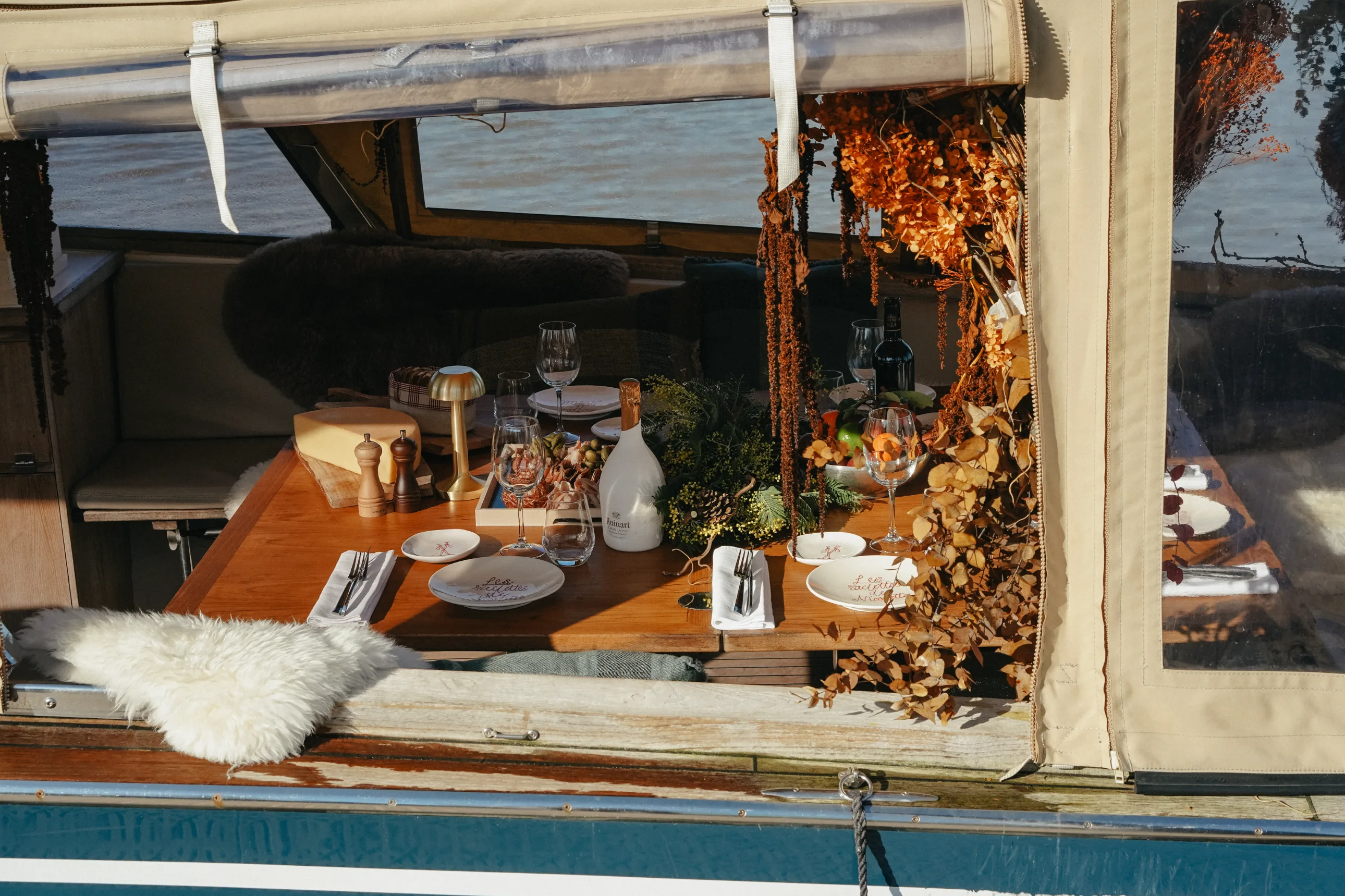Cozy dining table set for a meal inside a boat with plates, wine glasses, a cheese block, and autumnal floral decorations.