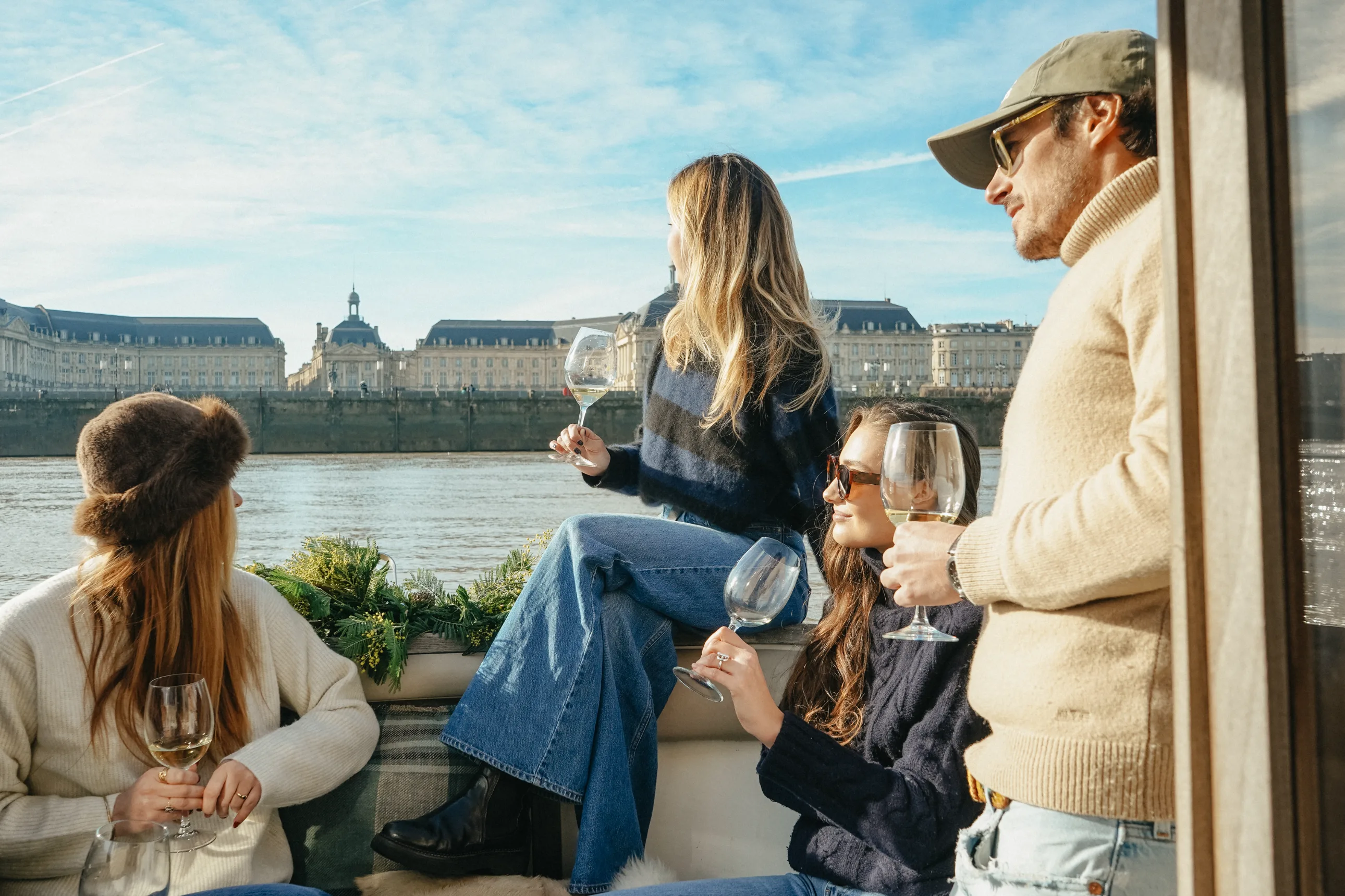 Four people sitting and standing on a boat by the river, holding wine glasses and enjoying a sunny day with historic buildings in the background.