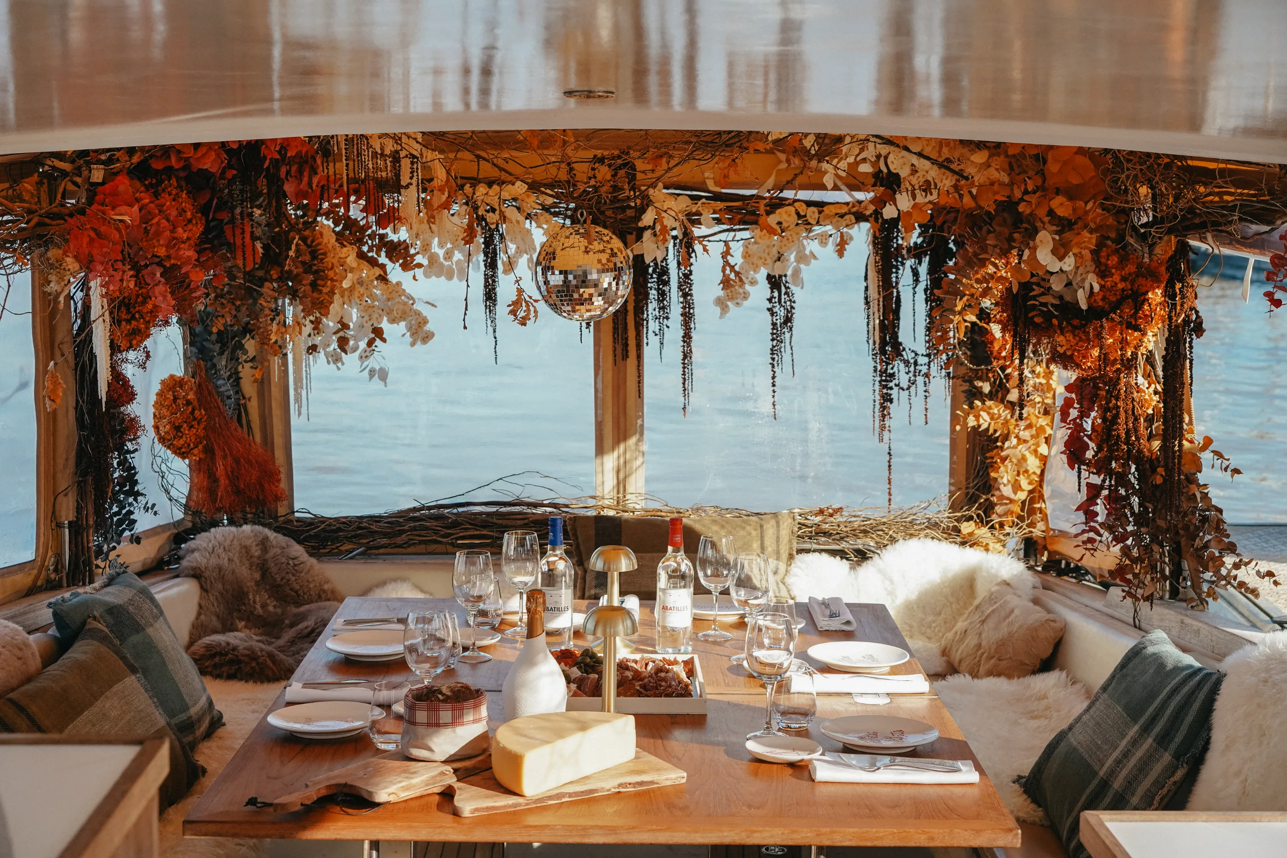 Cozy dining table set with wine bottles, cheese, and appetizers, surrounded by cushioned seating and autumn floral decorations by a water view.