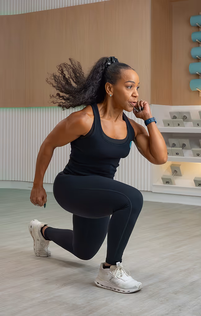 Woman in black athletic wear performing a forward lunge exercise indoors with her hair tied back and white sneakers.