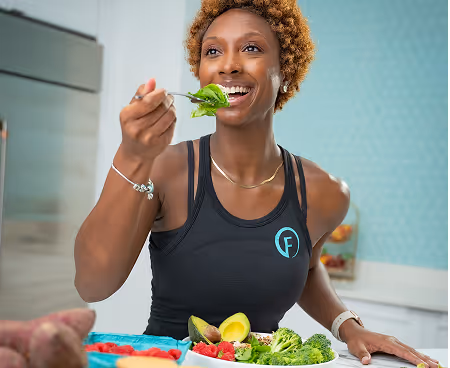 Smiling woman in athletic wear eating a healthy meal with broccoli, avocado, and strawberries in a kitchen.