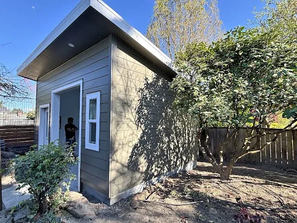 Gray-sided backyard building with a sloped roof, entry door, and narrow window