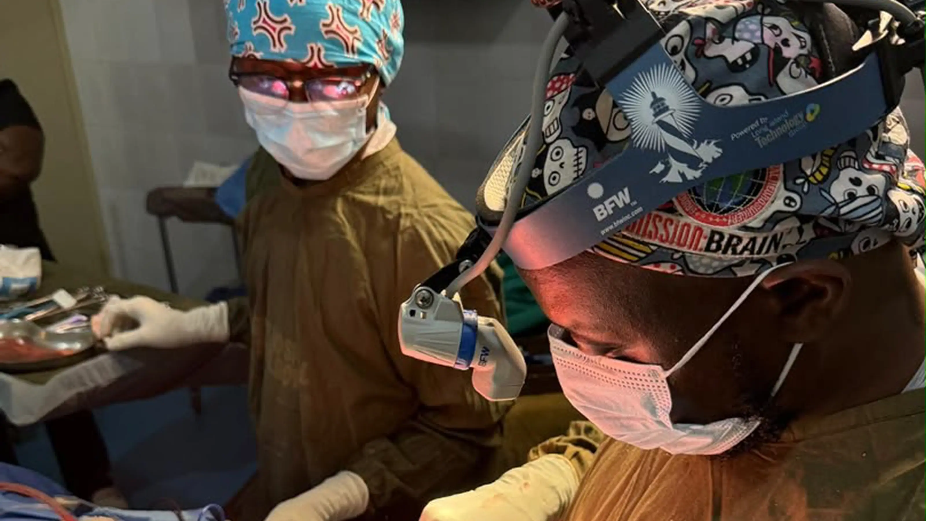 Dr. Alieu Kamara, wearing a mission:BRAIN surgical cap and headlight, performs Sierra Leone's first locally led brain tumor excision at Connaught Teaching Hospital in Freetown, with a masked colleague assisting beside the instrument table.