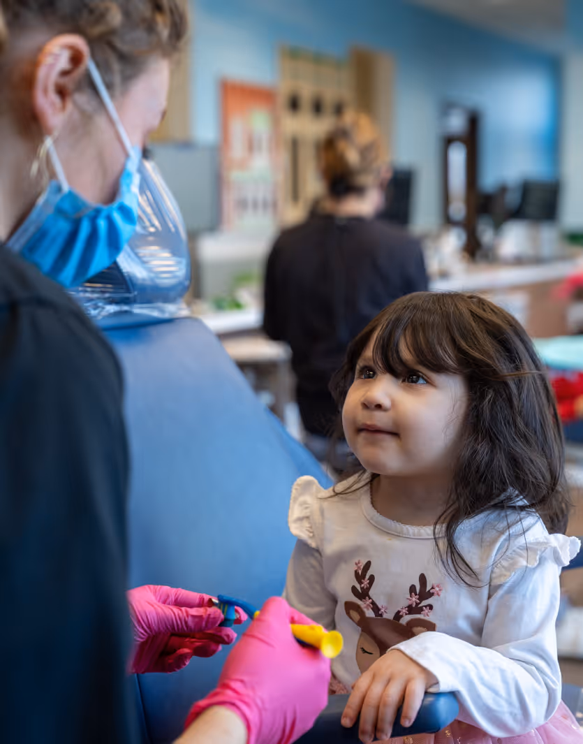 Child sitting on a dental chair looking at the dentist wearing a mask and pink gloves holding dental tools.