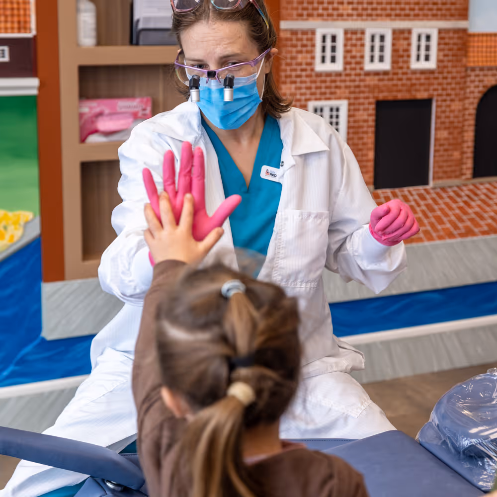 Dentist wearing a mask and magnifying glasses giving a pink-gloved high five to a child with a ponytail.
