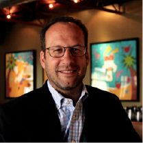 Smiling man with glasses and a checkered shirt under a black jacket in a warmly lit room with colorful artwork on the walls.