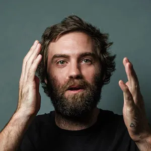 Bearded man with disheveled hair and a surprised expression, holding his hands near his face.