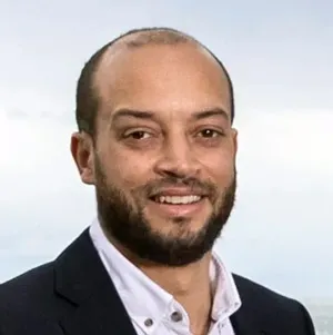 Smiling man with a beard and short hair wearing a white shirt and dark blazer against a light background.