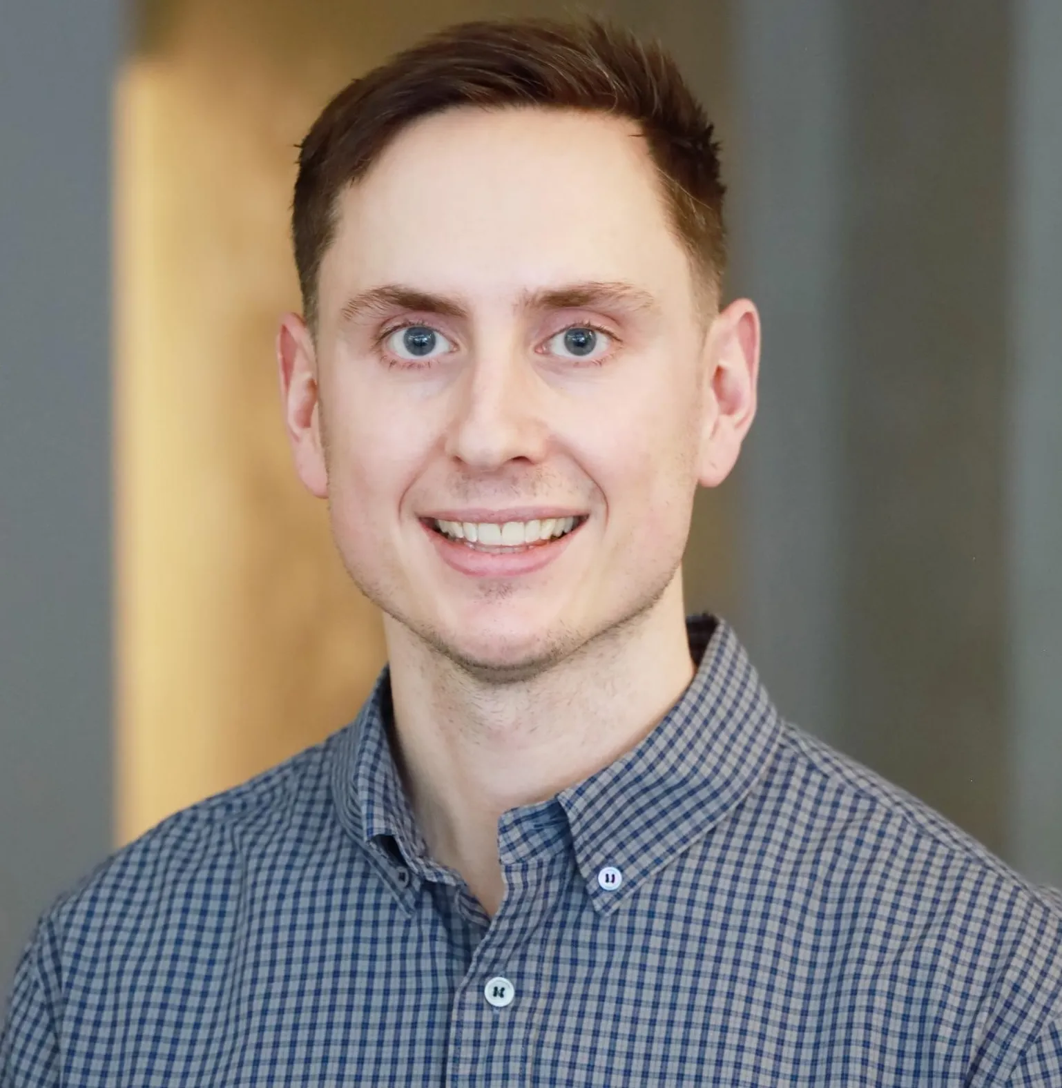Smiling young man with short brown hair wearing a blue checkered button-up shirt against a blurred background.
