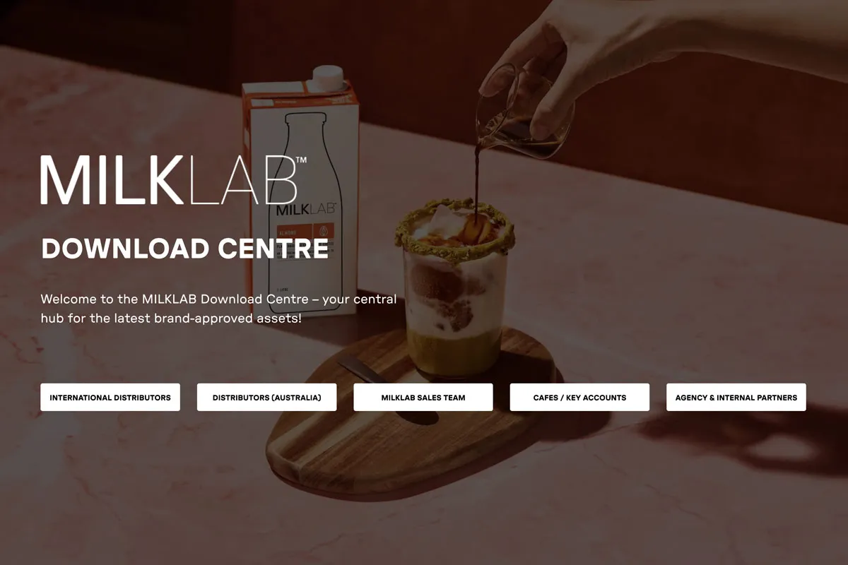 Hand pouring coffee into a glass with layered milk and a pistachio rim on a wooden board next to a MILKLAB carton on a marble surface.