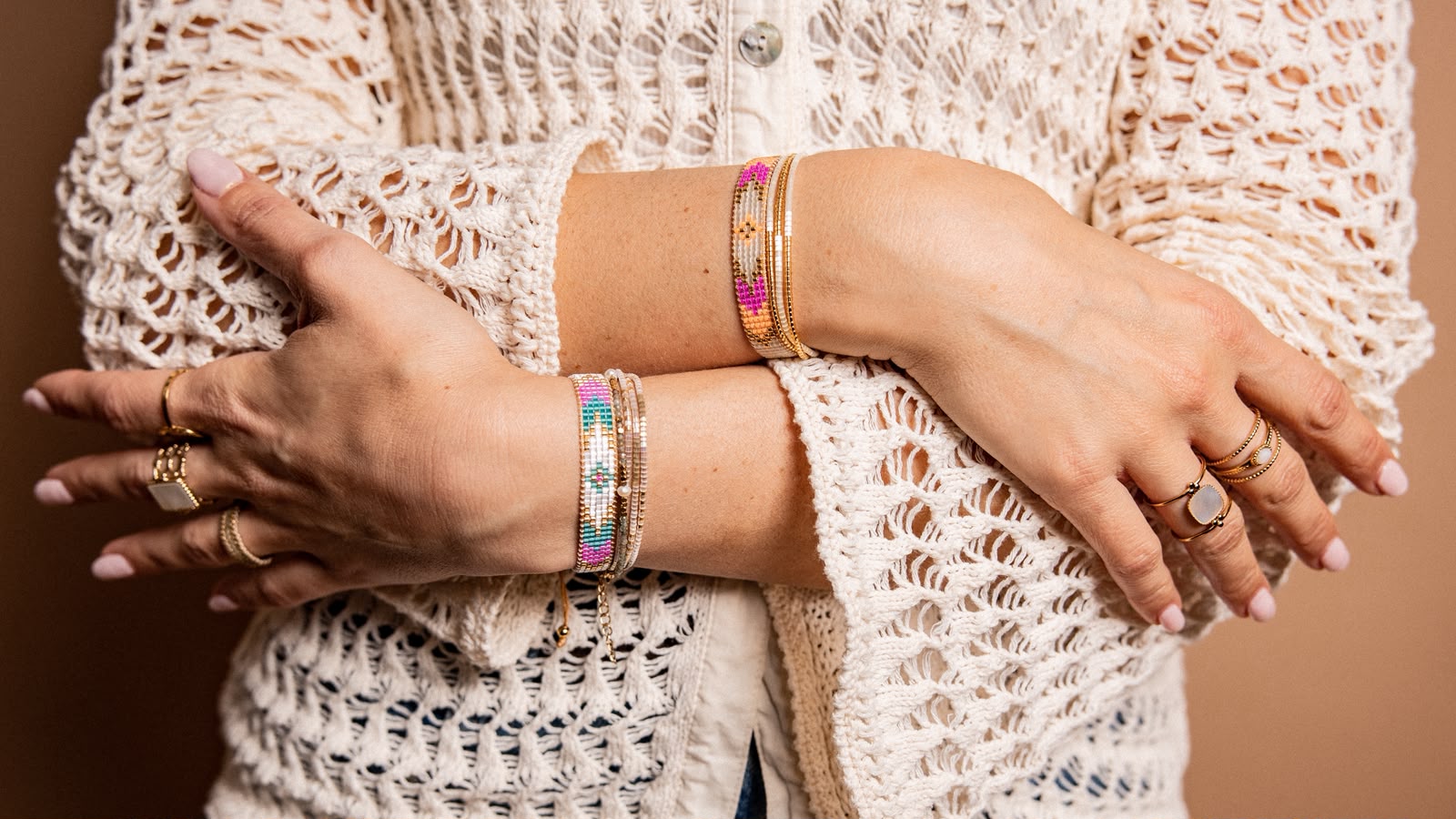Mains croisées d'une personne portant un pull beige en crochet et des bracelets et bagues colorés.