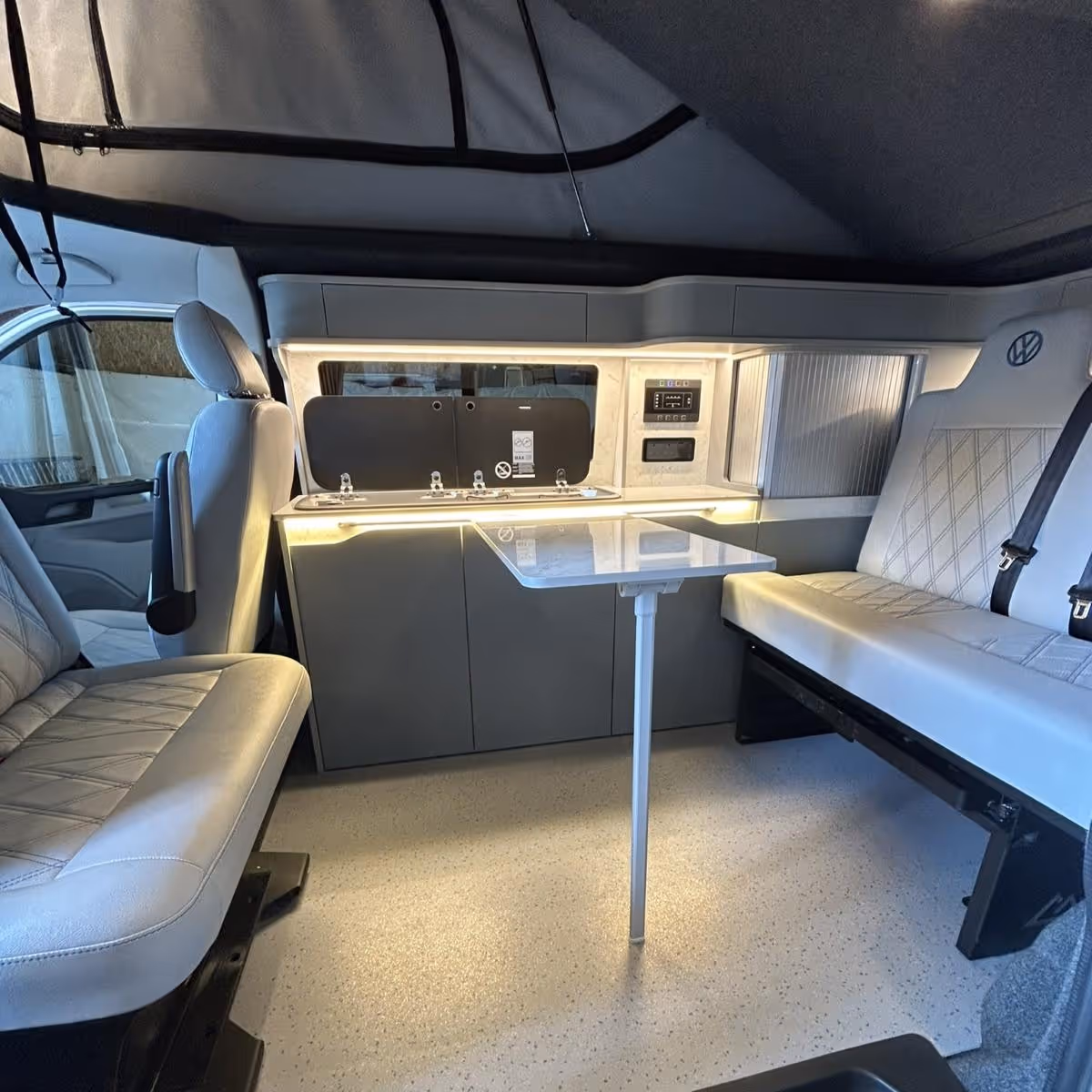 Interior of a camper van with two gray leather seats facing a small foldable table and a kitchenette equipped with a stove and overhead cabinets.