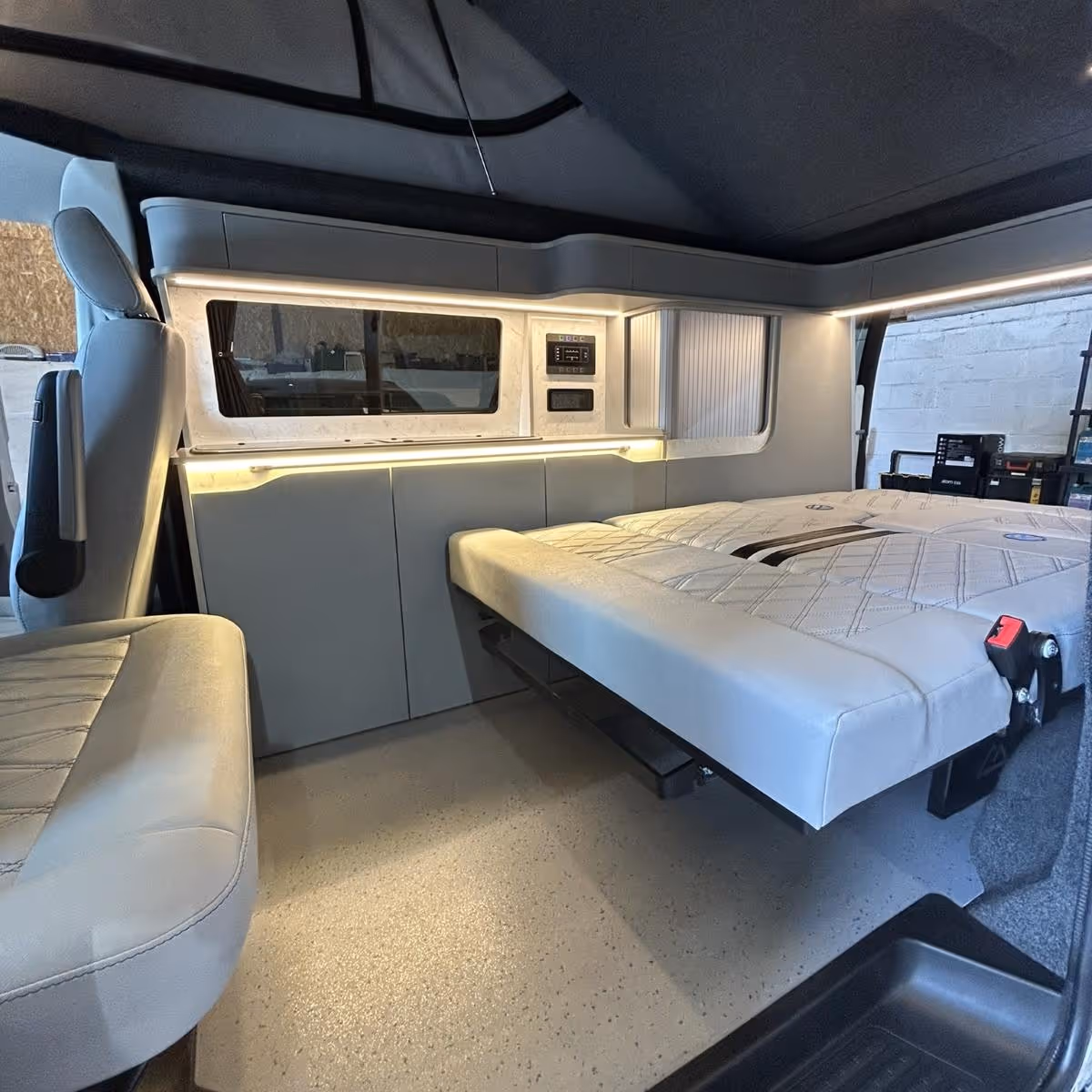 Interior of a camper van showing a fold-out bed next to a driver seat and storage cabinets with ambient lighting.