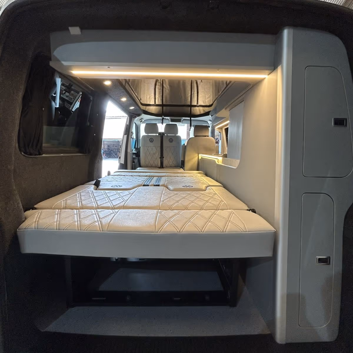 Interior rear view of a camper van converted with a beige quilted foldable bed and seating, illuminated by built-in strip and spotlights.