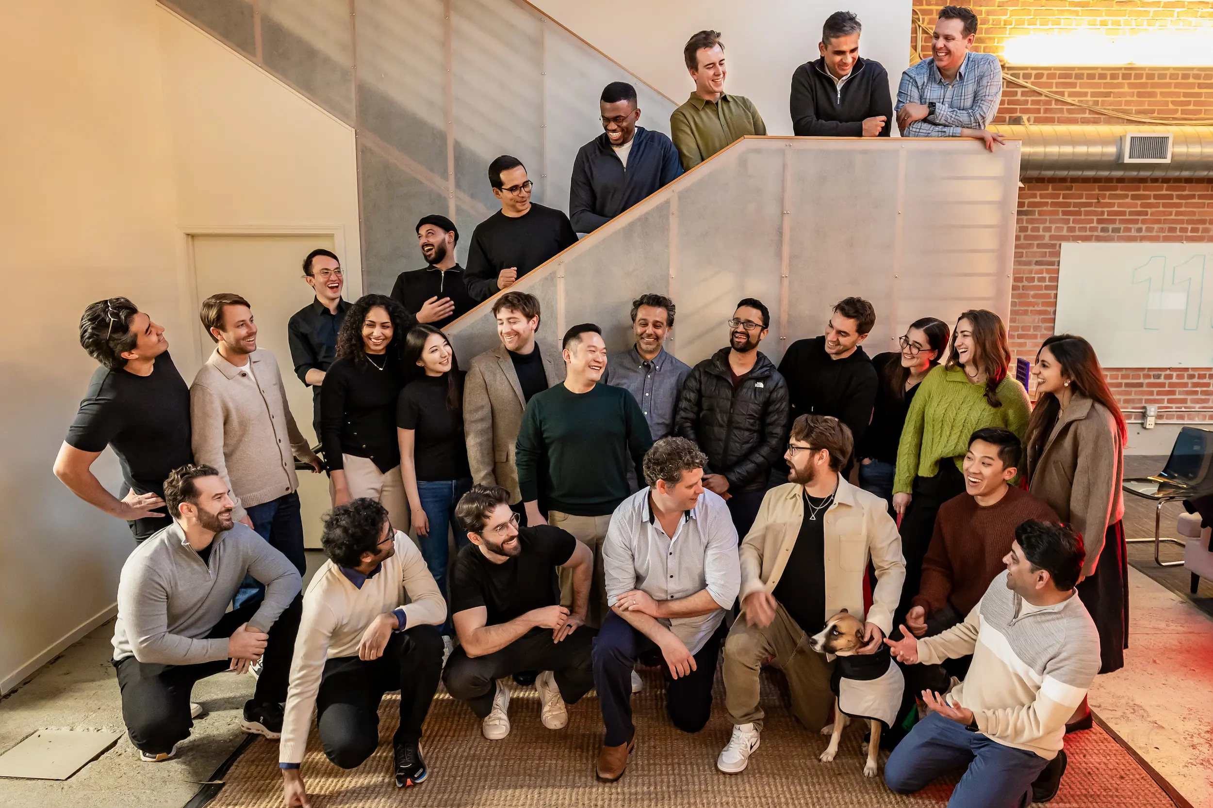 Group of diverse young adults posing together inside a modern office space, some standing and some crouching, with a dog wearing a coat.