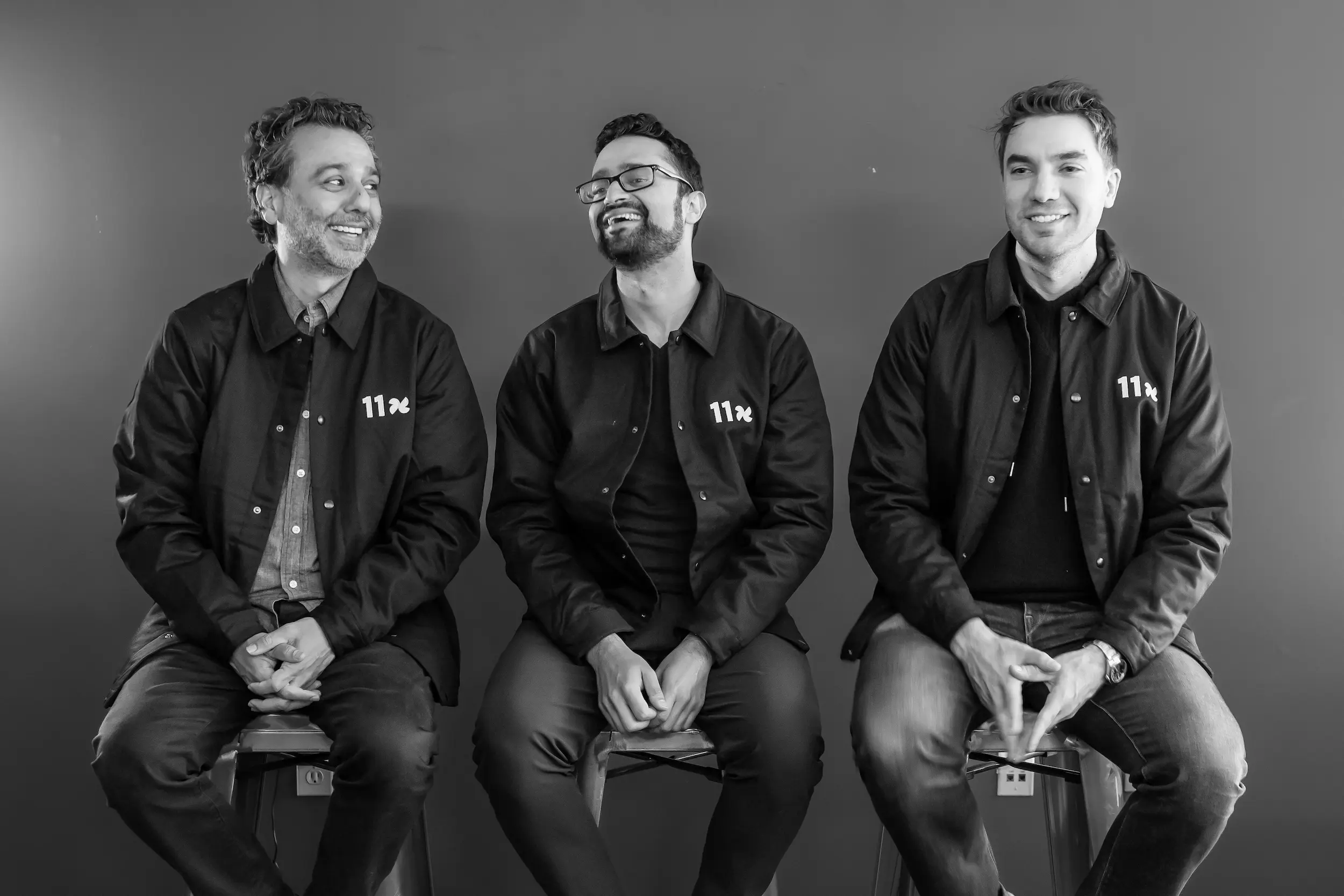 Three men sitting on stools against a plain wall, wearing matching jackets with '11x' logos, two smiling and one looking to the side.