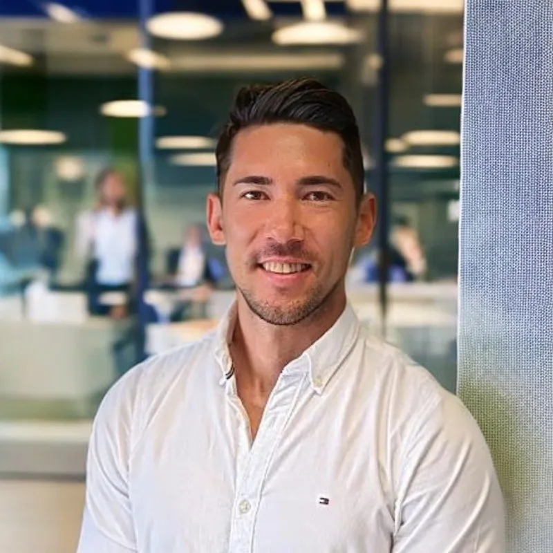 Smiling man with short dark hair wearing a white button-up shirt standing in an office with blurred coworkers in the background.