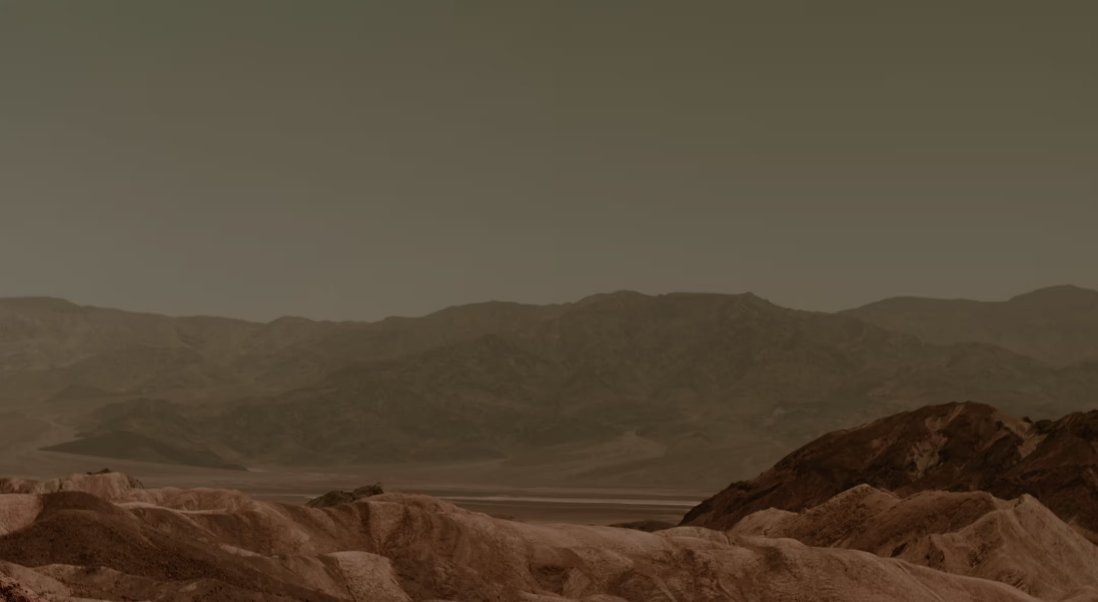 Desert landscape with rocky hills in the foreground and distant mountains under a muted sky.