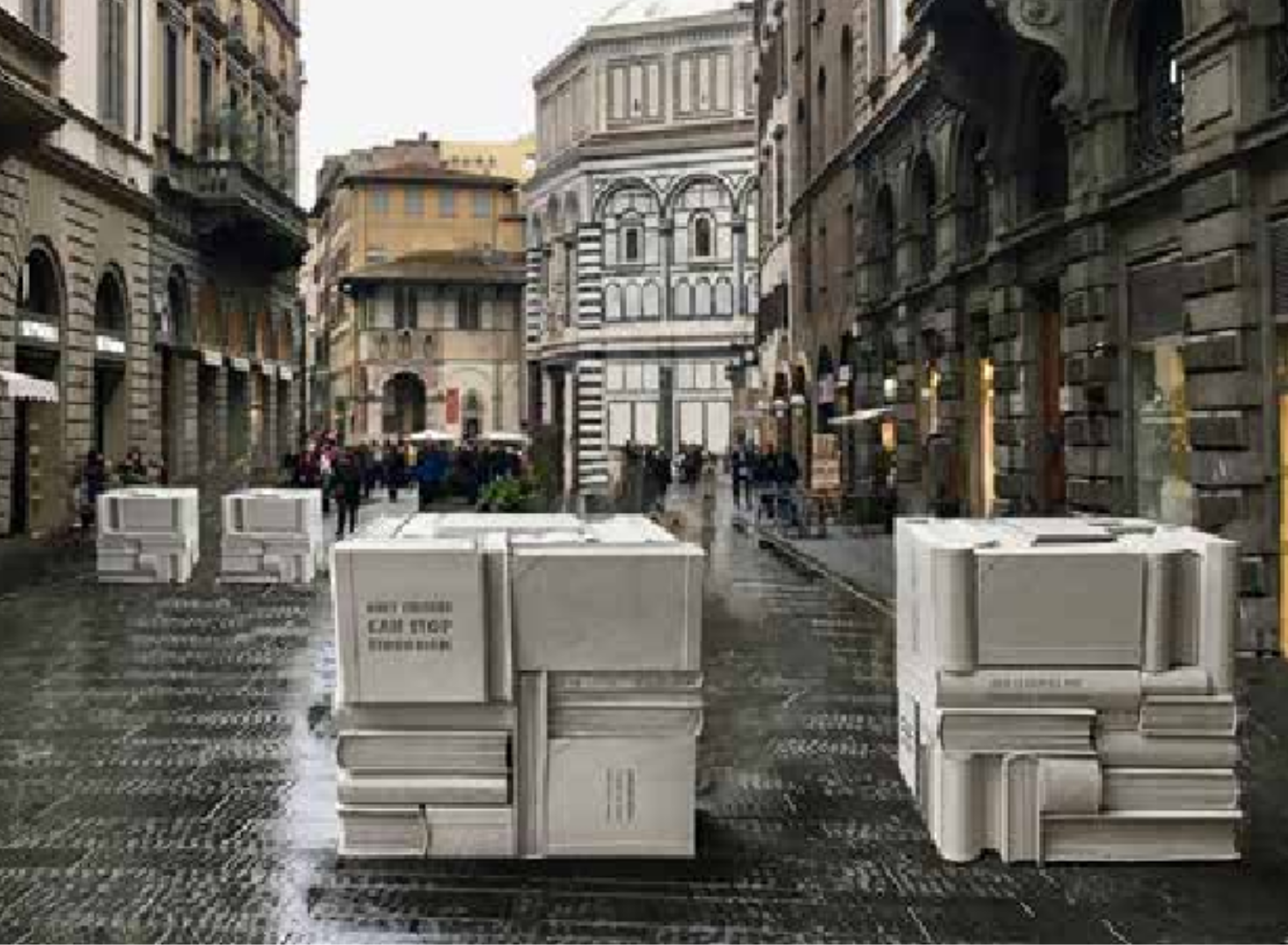 Scultura a forma di libri impilati in una piazza acciottolata con edifici storici sullo sfondo e persone che camminano.