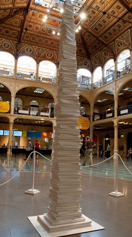 Una torre alta composta da grandi pile di libri bianchi esposti in un grande atrio con soffitto decorato.