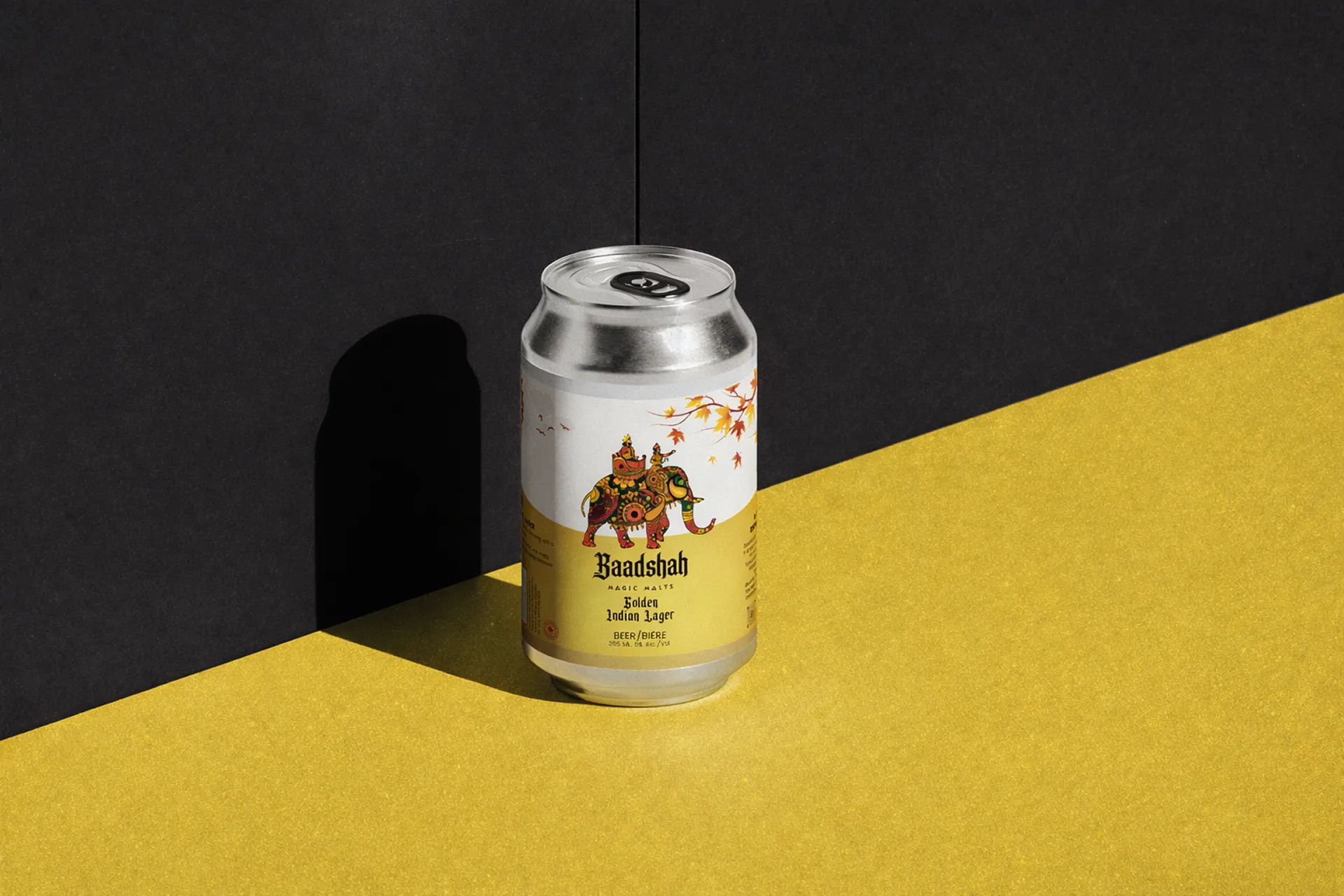 Silver can of Baadshah Golden Indian Lager beer with colorful decorated elephant artwork on the label, set against a two-tone black and yellow background casting a shadow.
